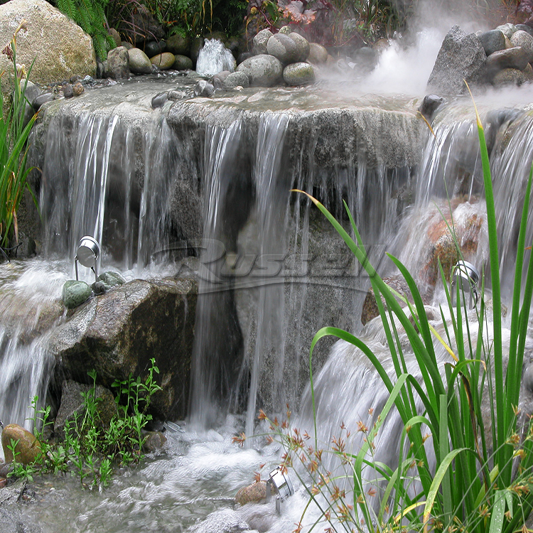 A natural-looking cascading waterfall feature with layered rock tiers, flowing water, mossy stones, and tall green reeds in the foreground.